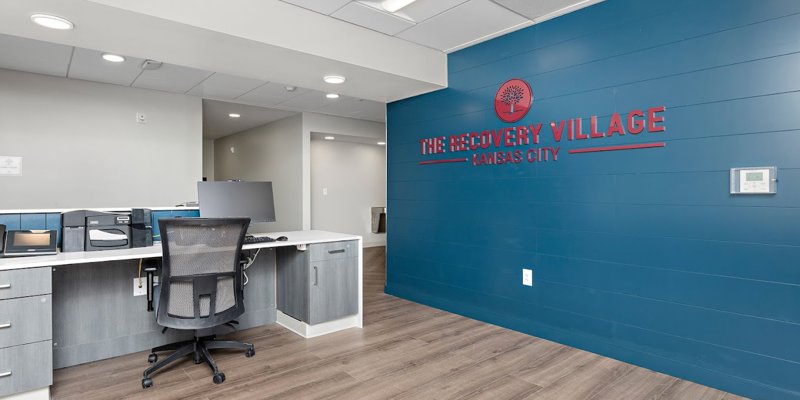 A clean and modern reception area with sleek furniture, wood flooring, and a bold blue wall displaying the "Recovery Village Kansas City" logo.