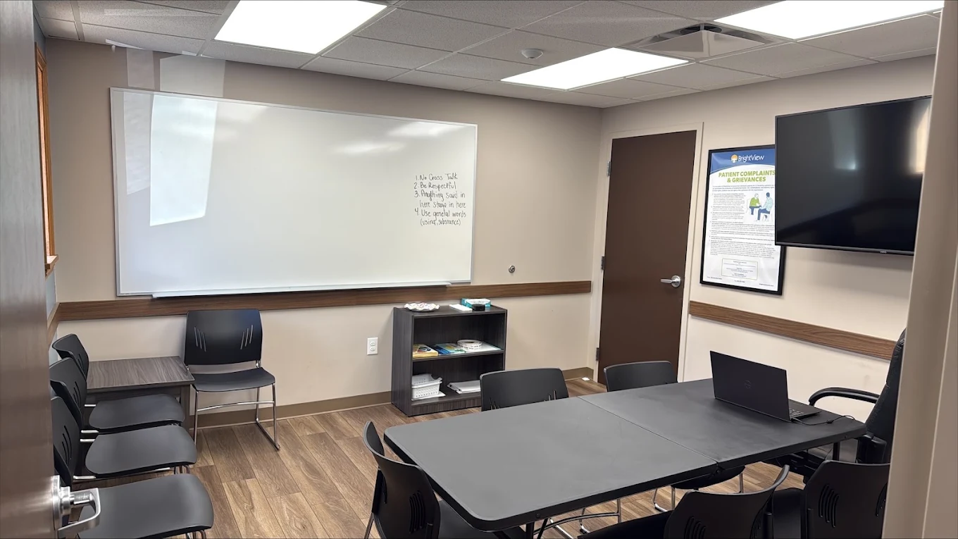 Group therapy room with conference table, black chairs, whiteboard, wall-mounted television, and patient information poster