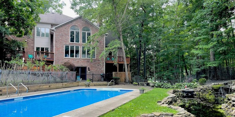 Serene backyard with a sparkling pool, lush greenery, a tranquil pond with a waterfall, and a charming brick house surrounded by tall trees.