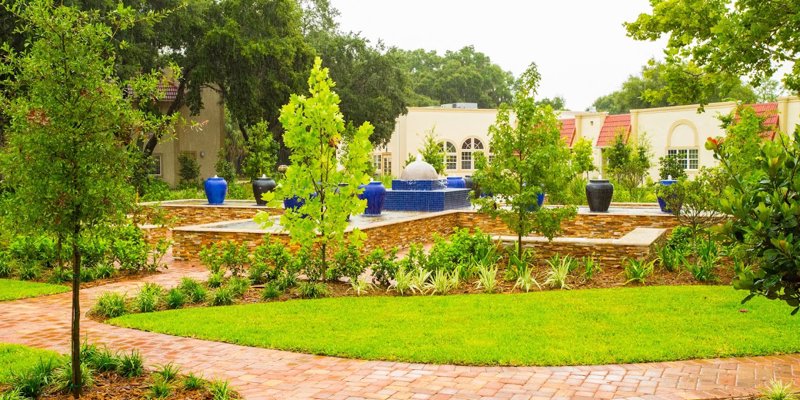 A serene courtyard featuring lush greenery, vibrant blue vases, and a central water fountain surrounded by peaceful pathways and gardens.