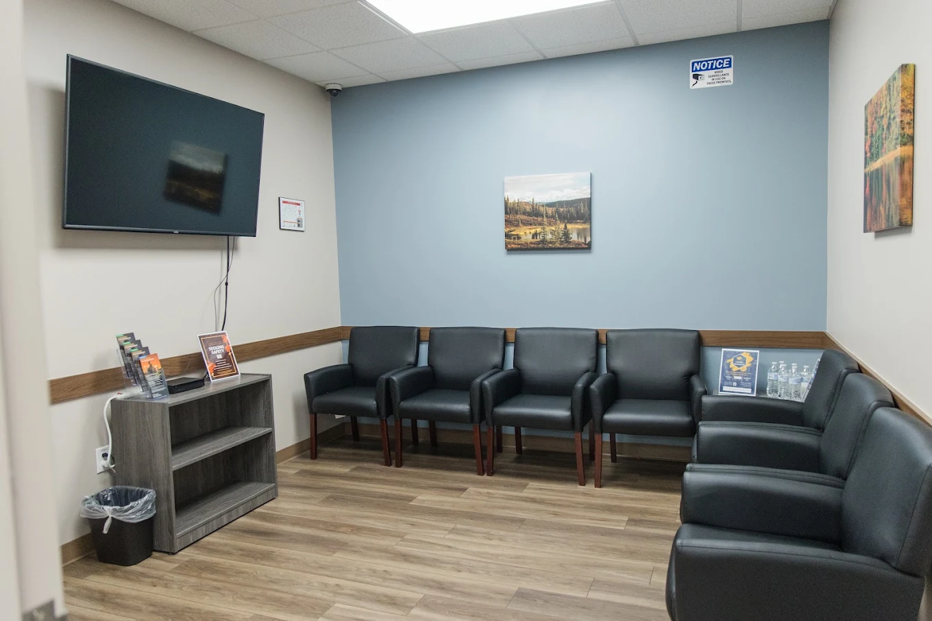 Treatment waiting room with black armchairs, wall-mounted television, informational materials, and landscape artwork