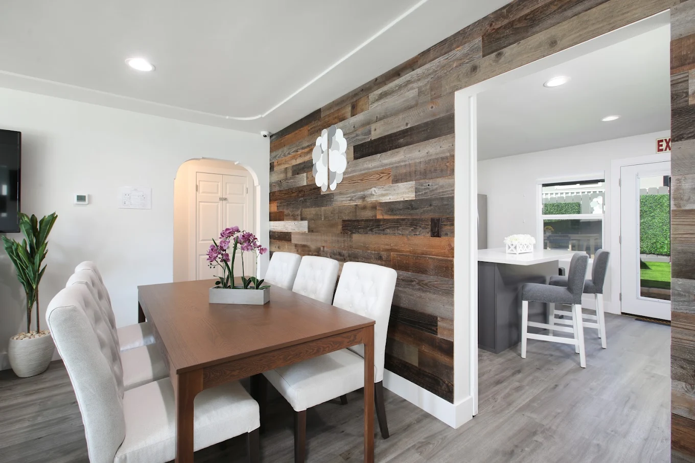 Dining area with wood table, white upholstered chairs, reclaimed wood accent wall, and adjacent kitchen