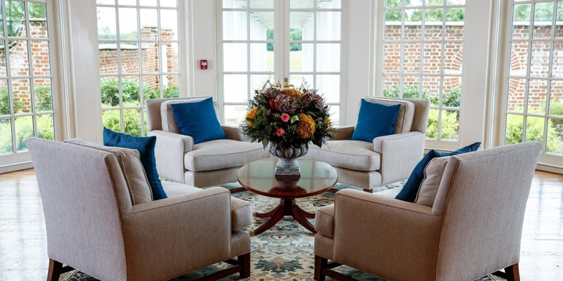 Bright sunroom with cozy armchairs, blue accent pillows, a central floral arrangement, and large windows offering views of a charming garden.