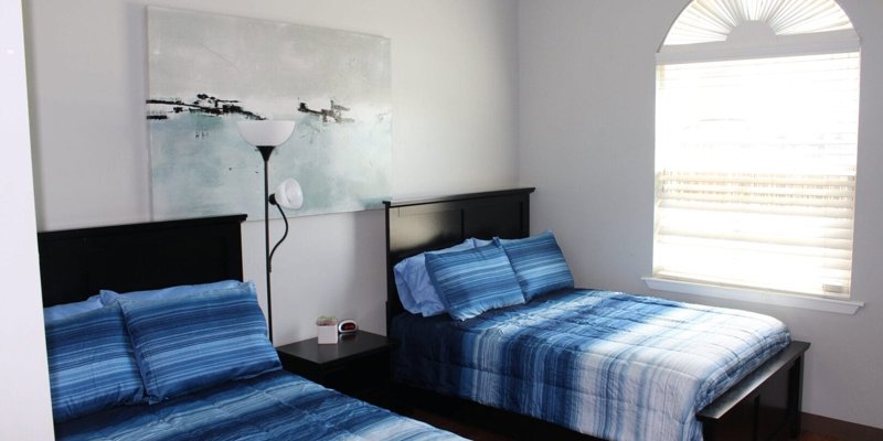  A bright and cozy bedroom with two neatly made beds featuring blue bedding, a large window letting in natural light, and modern wall art.