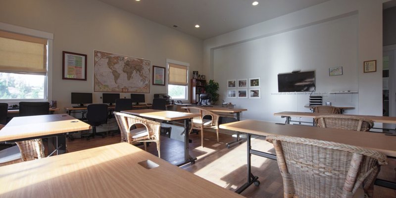 A spacious classroom with large windows offers plenty of natural light, featuring wicker chairs and modern desks for a comfortable learning environment.