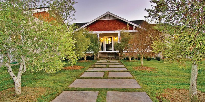 A beautifully landscaped walkway leads to a charming, warmly lit home, framed by lush trees and a well-maintained lawn, offering a welcoming ambiance.