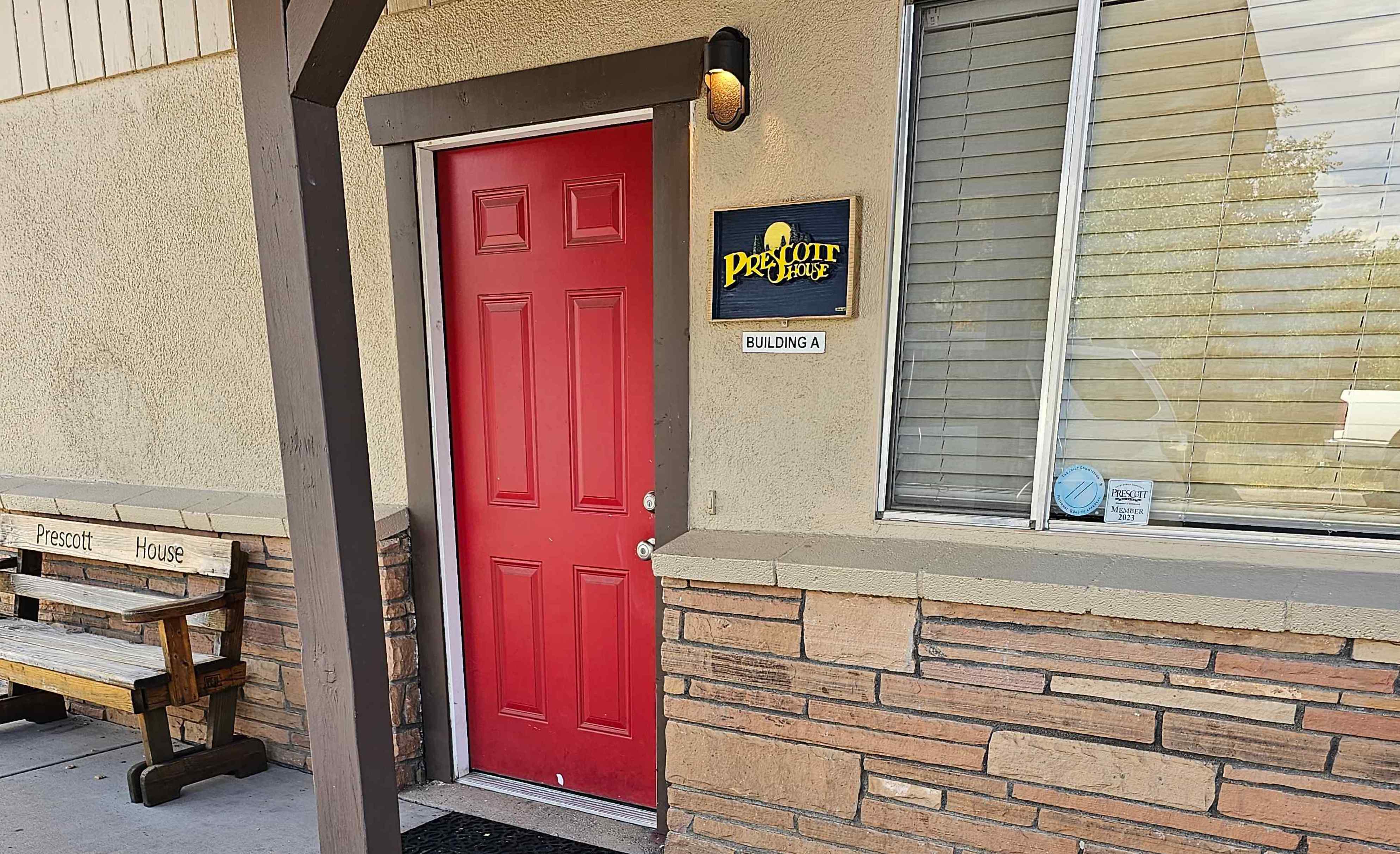 Entrance with red door, Prescott House sign, bench, and stone-trimmed beige walls.