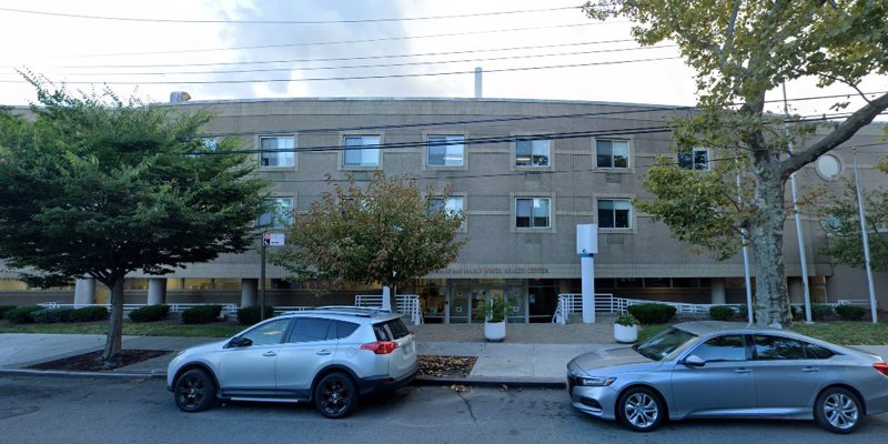 A modern building with clean lines, shaded by large trees and framed by parked cars, creating a calm and well-maintained urban setting.