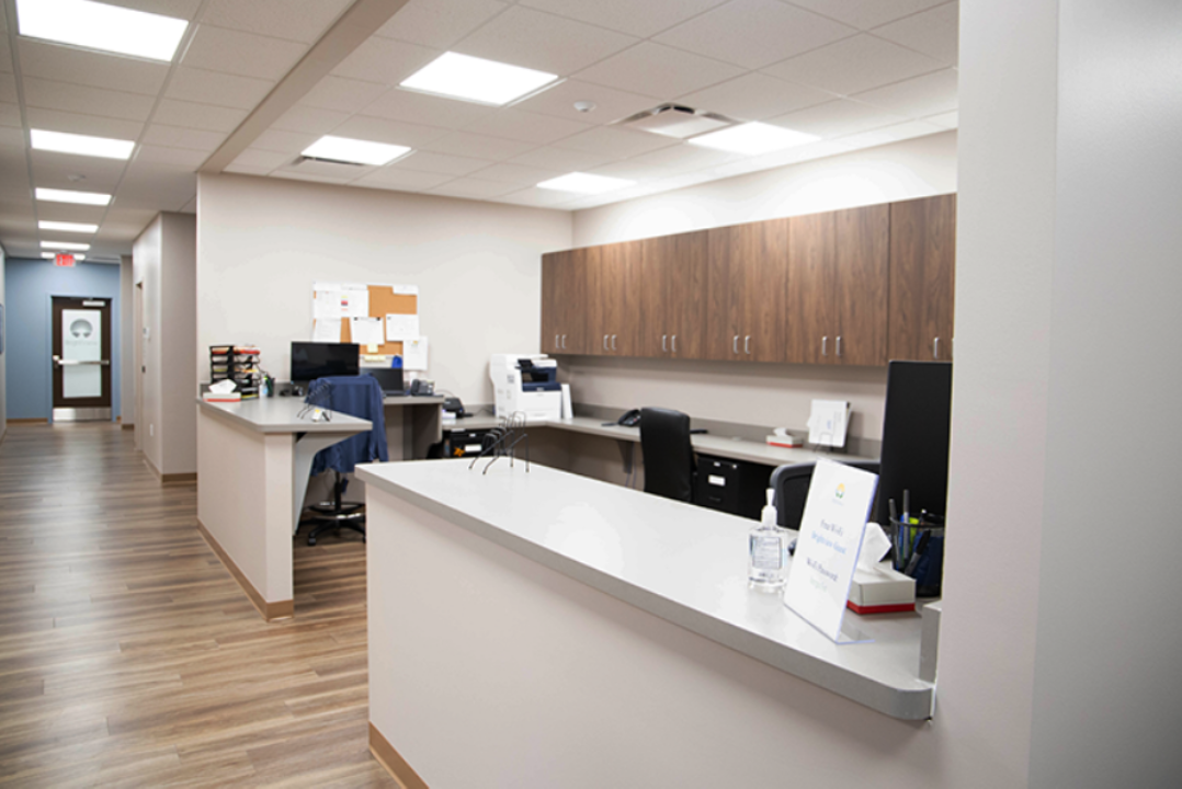 Staff workstation with reception counters, computers, office equipment, overhead wood cabinets, and adjacent hallway