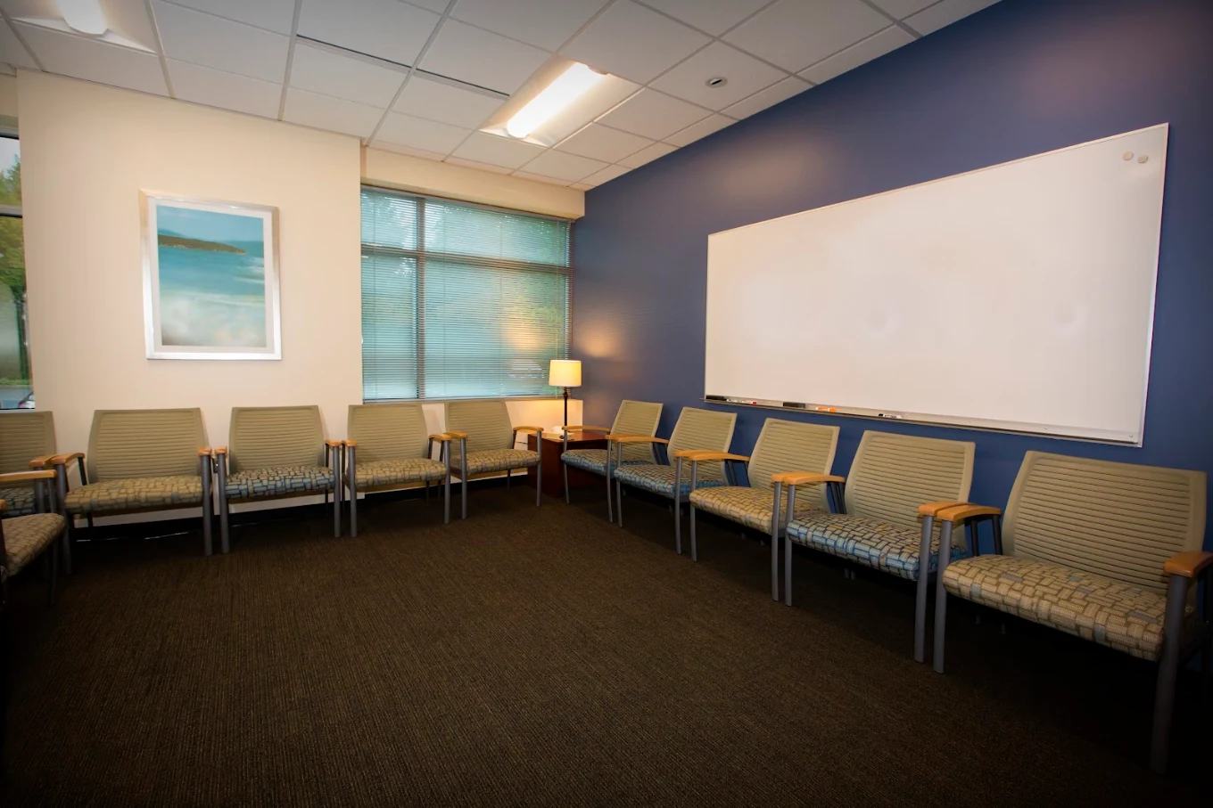 Therapy room with chairs arranged around the walls, a large whiteboard, windows, and soft lighting
