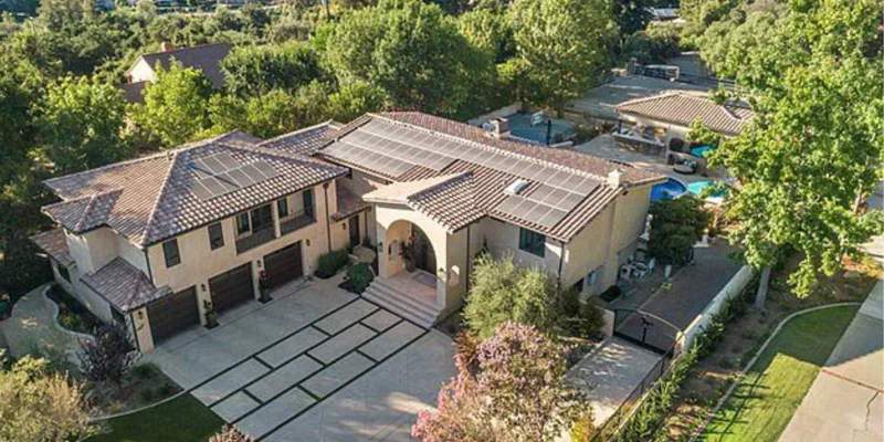 Aerial view of a beautiful, expansive home with solar panels, surrounded by lush greenery, nestled in a serene, tree-filled neighborhood.