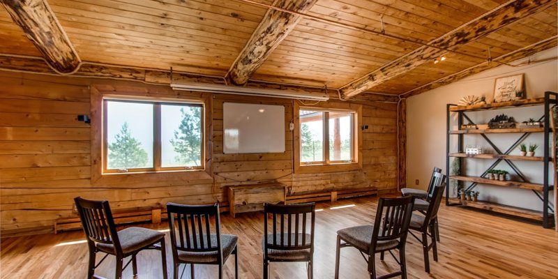 A rustic meeting room with wooden walls, exposed beams, large windows with scenic views, and chairs arranged for a group session, creating a warm and inviting space.