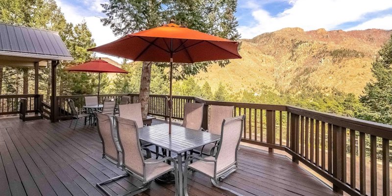 A spacious wooden deck with outdoor dining tables, shaded by orange umbrellas, overlooking scenic mountain views and surrounded by trees.