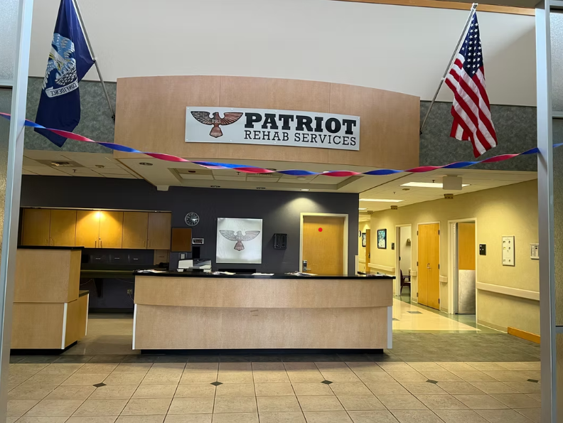 Reception desk with a sign reading “Patriot Rehab Services,” U.S. flags, and hallways leading to treatment areas