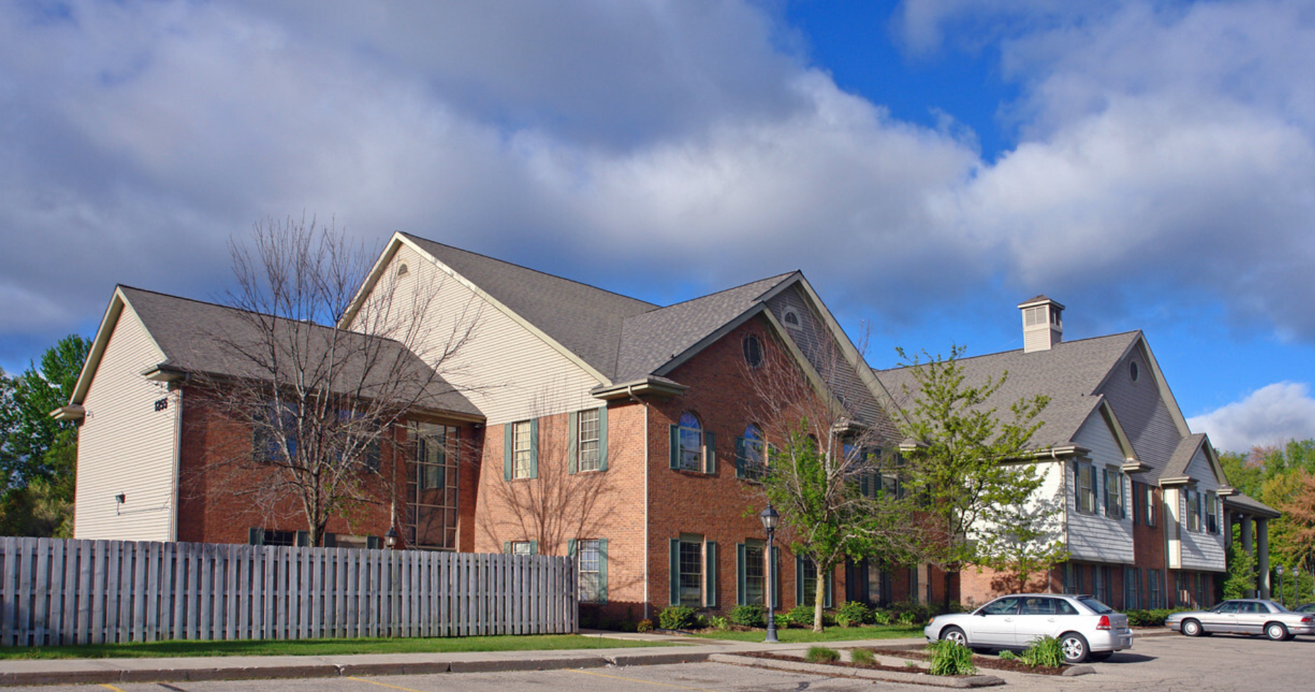 A large two-story building with a combination of red brick and white siding, featuring multiple windows, a pitched roof, and a parking lot in front, serving as the Community Programs Inc. facility.