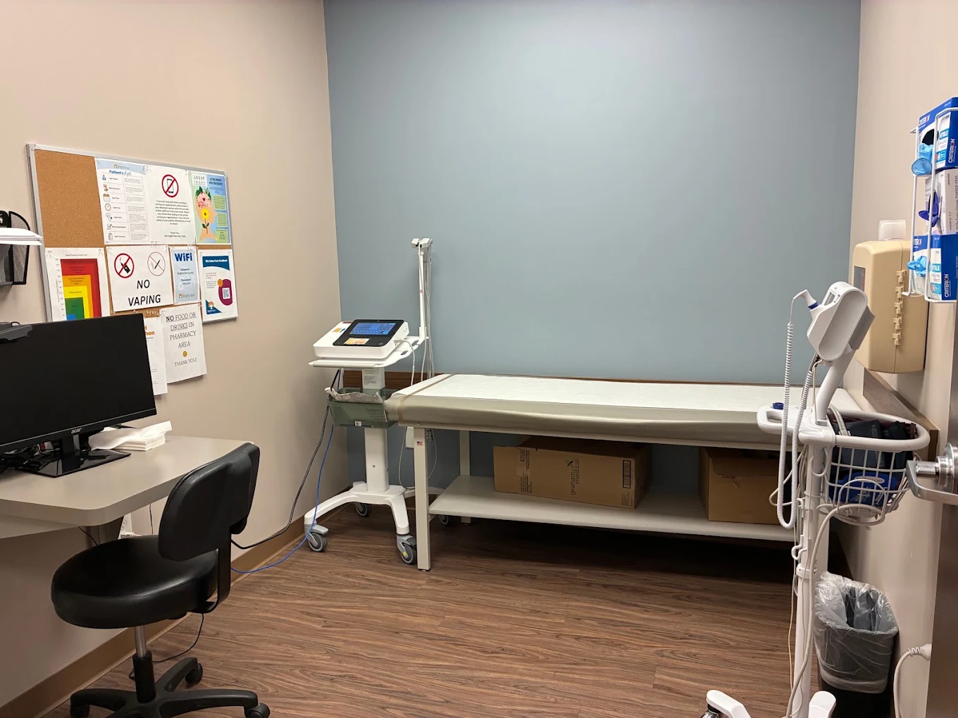 Medical exam room with examination table, computer workstation, vital-sign equipment, clinical supplies, and informational notices