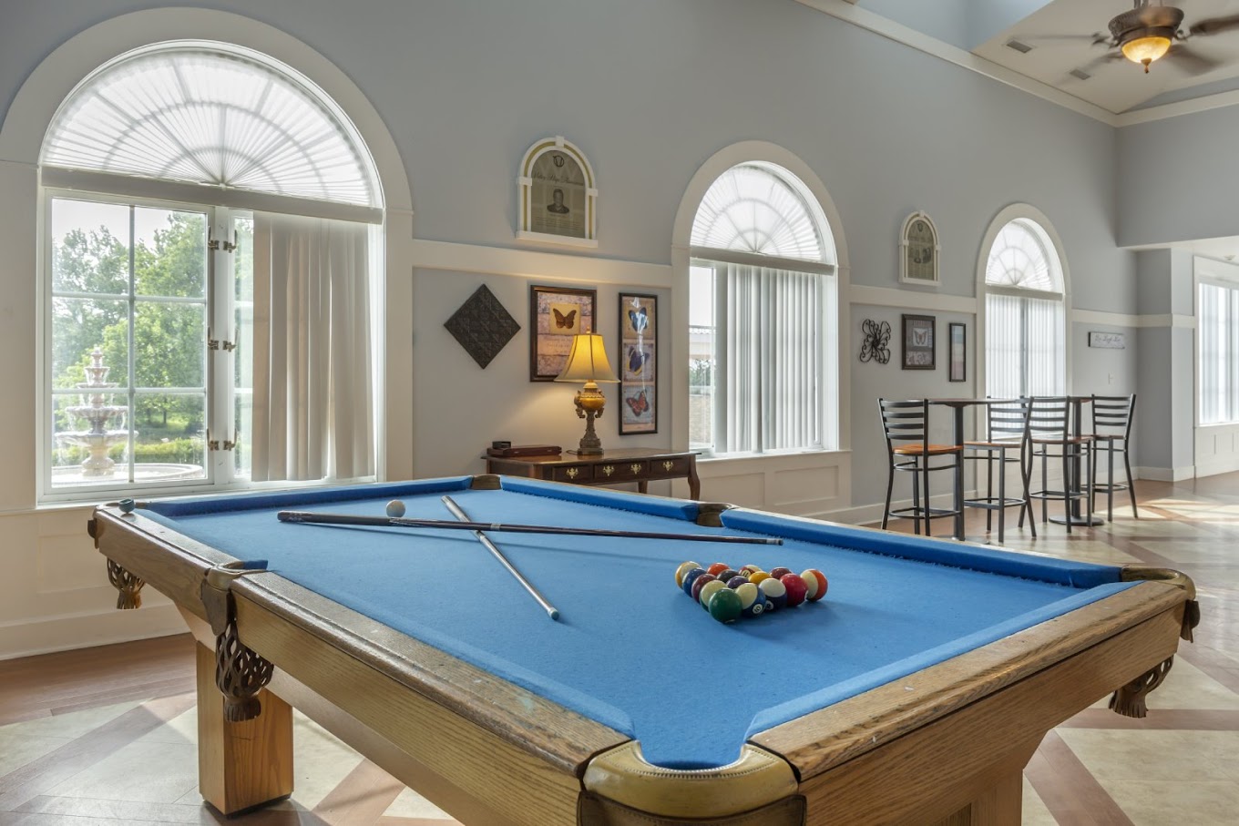 A well-lit game room with a blue pool table, classic decor, and arched windows offering a scenic view.