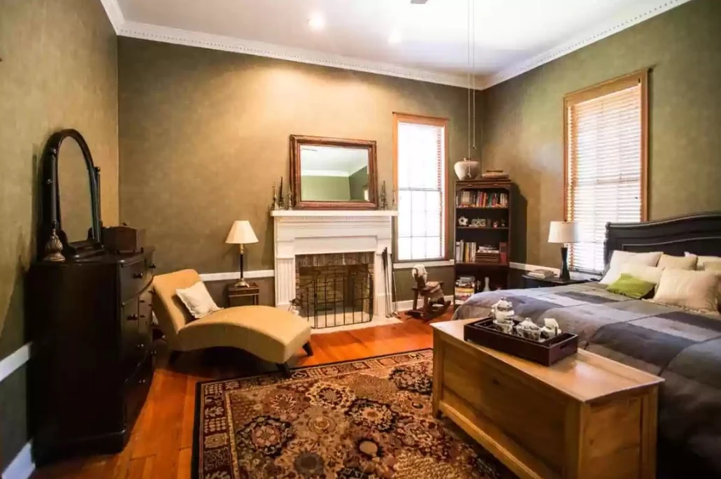 Traditional bedroom with large bed, fireplace, chaise lounge, wood flooring, dresser, bookshelf, and patterned area rug