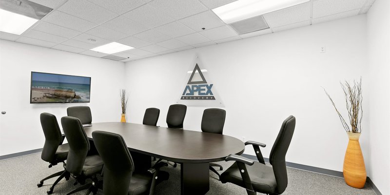 A modern conference room with a sleek table, black chairs, and a wall-mounted screen. The “Apex Recovery” logo is prominently displayed.