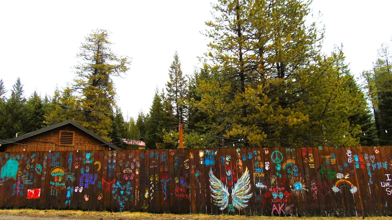 Colorful handprints, artwork, and messages painted on a wooden wall surrounded by evergreen trees at the Columbia Falls campus