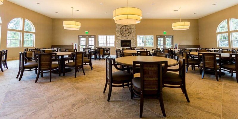 A spacious dining area with round wooden tables and comfortable chairs, illuminated by elegant ceiling lights and large arched windows for natural light.