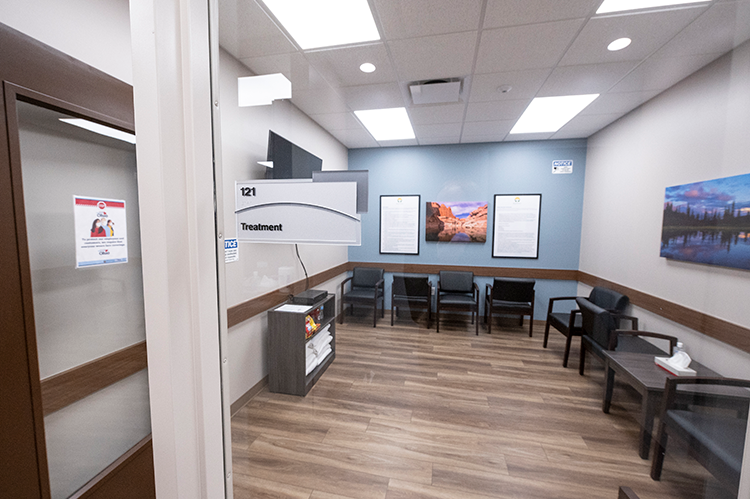 Treatment room with black patient chairs arranged along the walls, small tables, framed artwork, television, and wood-look flooring
