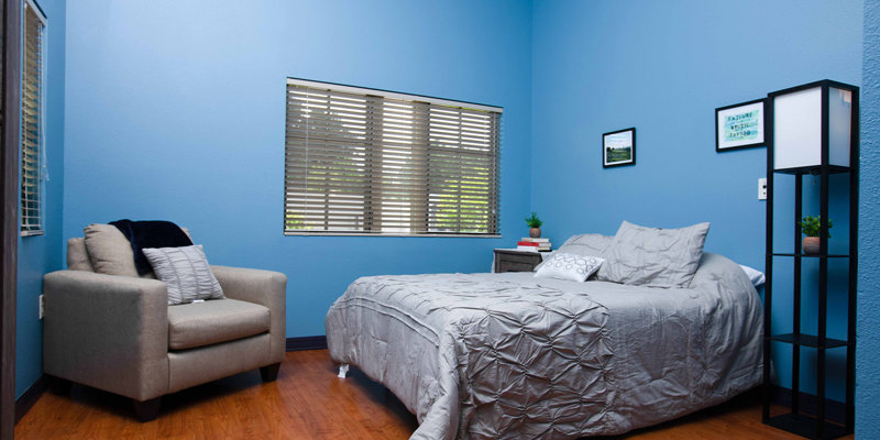 A cozy bedroom with calming blue walls, a comfortable bed, an armchair, and natural light from the window, offering a peaceful and relaxing space.