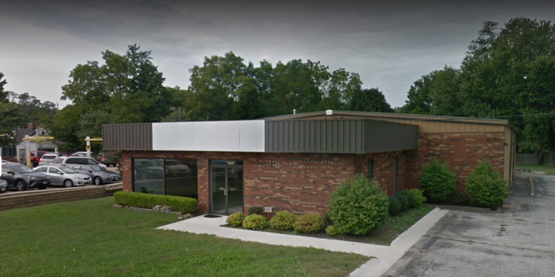 A brick office building with large windows and neatly trimmed bushes in front, surrounded by greenery and a small parking area.