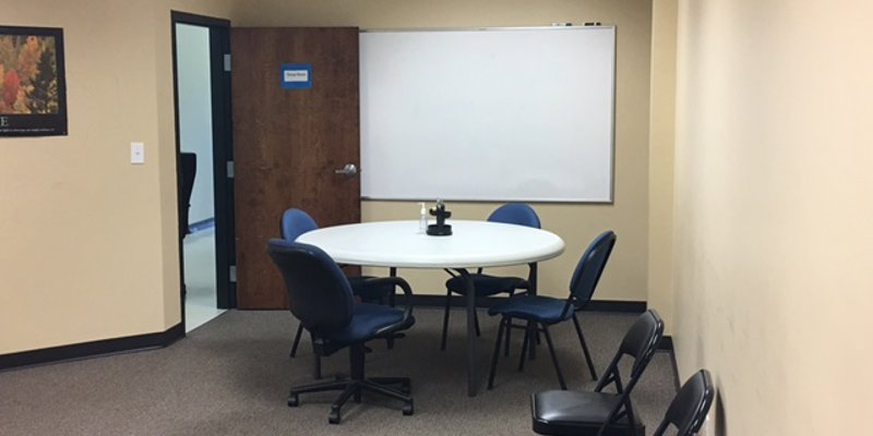 A small meeting room with beige walls, a round table surrounded by blue chairs, and a whiteboard, offering a calm, collaborative space.