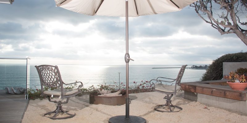 Tranquil outdoor seating area with an umbrella and ocean views, featuring sand underfoot and a cozy fire pit for relaxed gatherings.