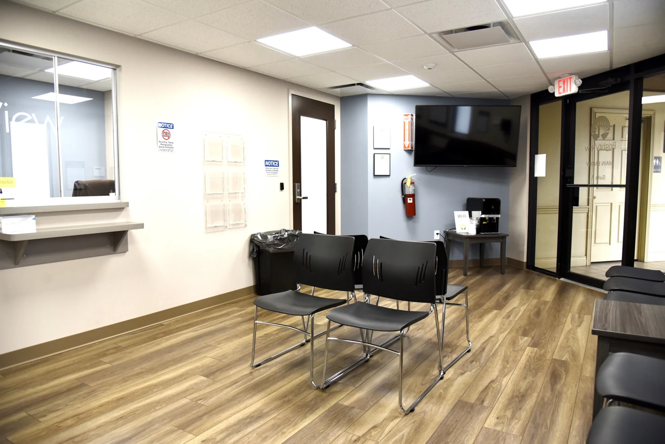 BrightView Health reception and waiting area with black chairs, service window, wall-mounted television, coffee station, wood-look flooring, and glass entrance doors