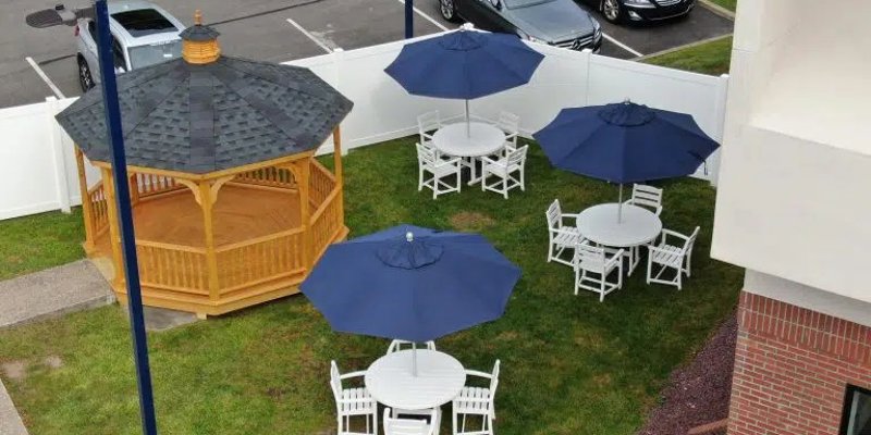 A peaceful outdoor space featuring a wooden gazebo, white patio tables with blue umbrellas, and a well-maintained grassy area for relaxation.