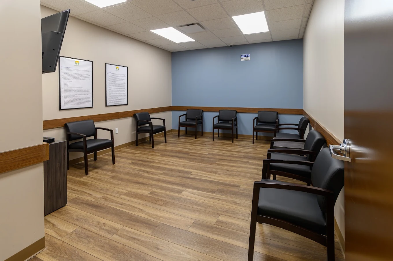 BrightView Health group treatment room with black cushioned chairs arranged around an open floor, blue accent wall, framed notices, television, and wood-look flooring