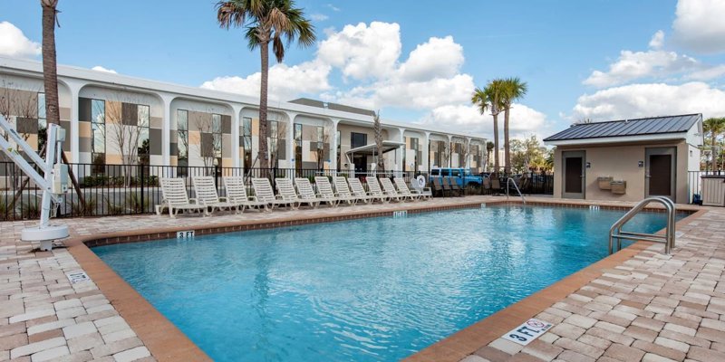 A sparkling outdoor pool surrounded by lounge chairs, set against a backdrop of palm trees and a modern building, offers a relaxing retreat.