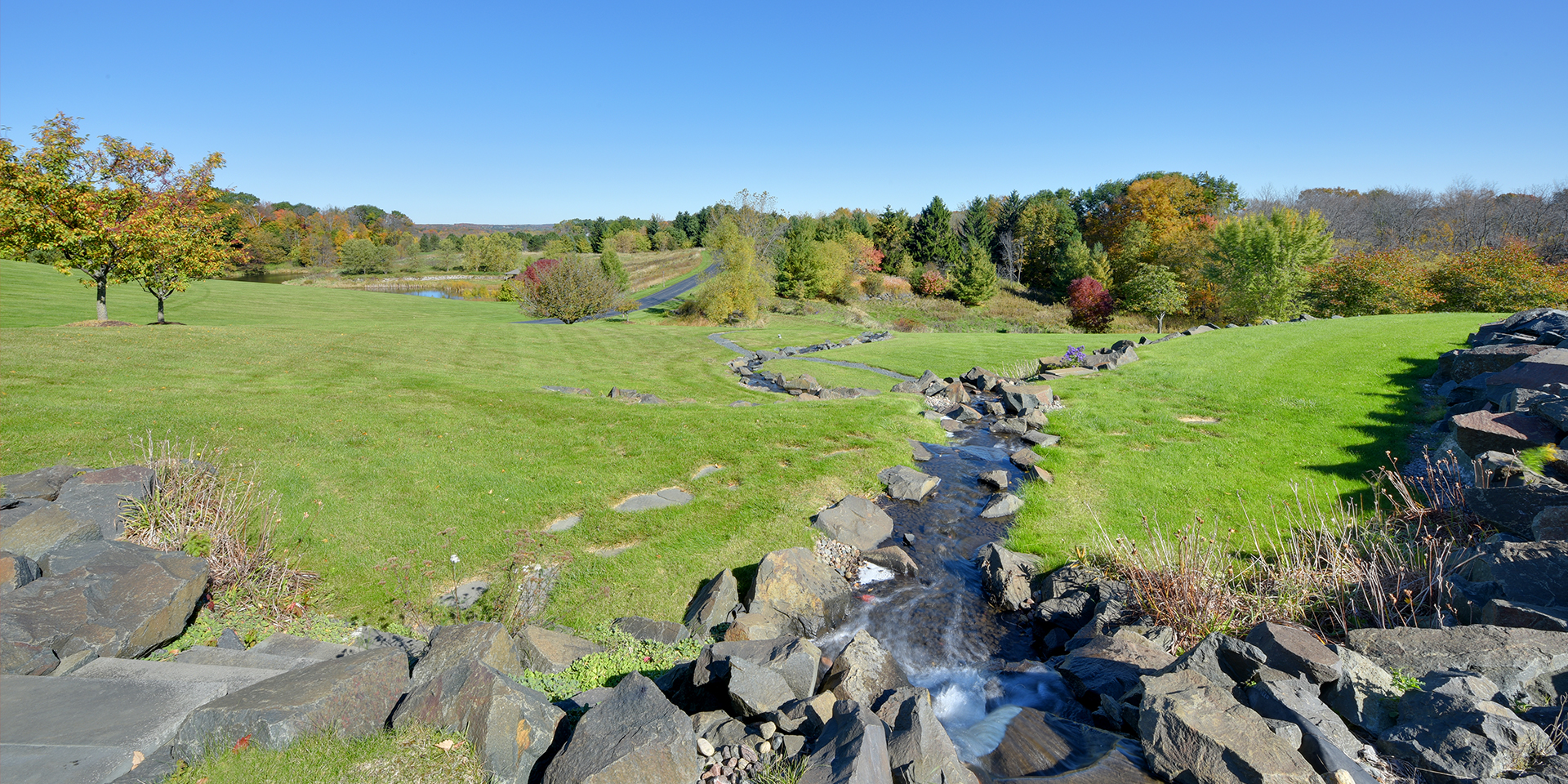 Stream flowing through landscaped lawn with rocks and autumn trees.