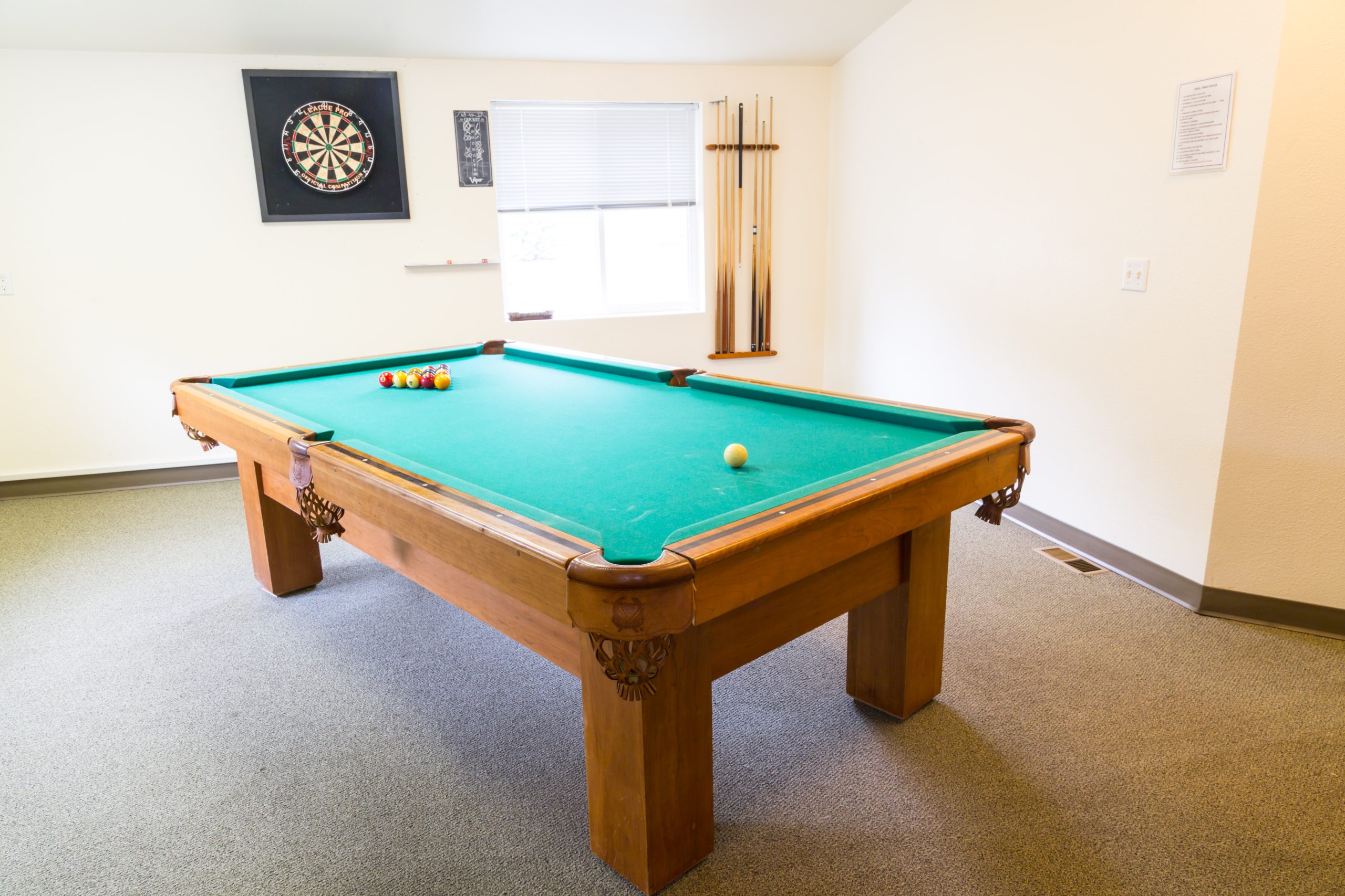 A pool table in a recreational room with a dartboard and cue sticks.
