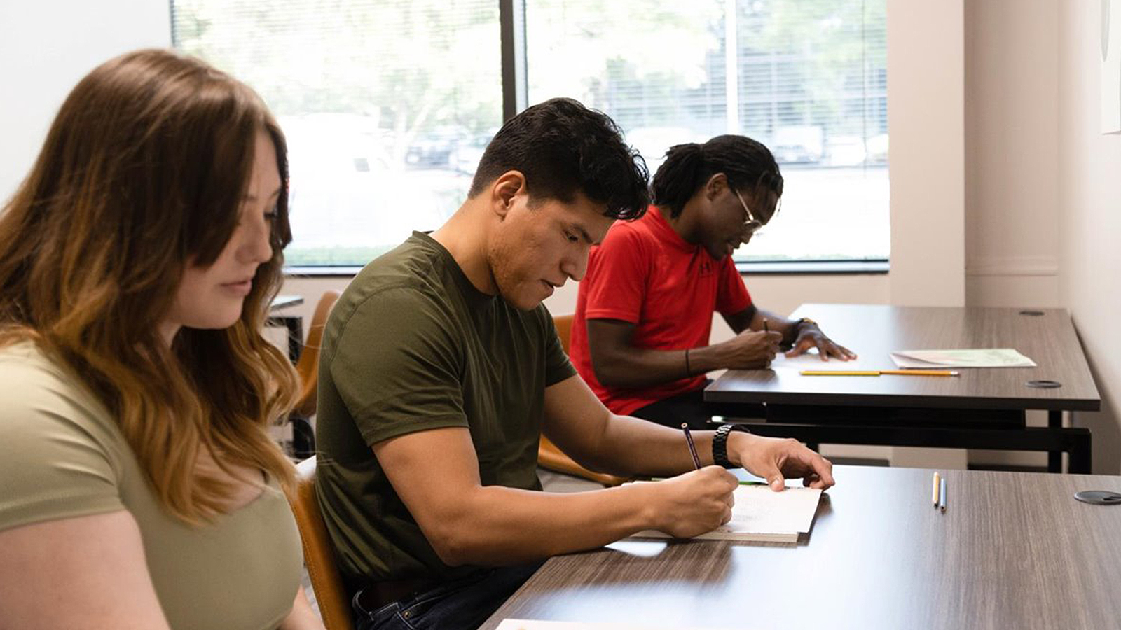 Three people writing at wooden desks in a bright room.
