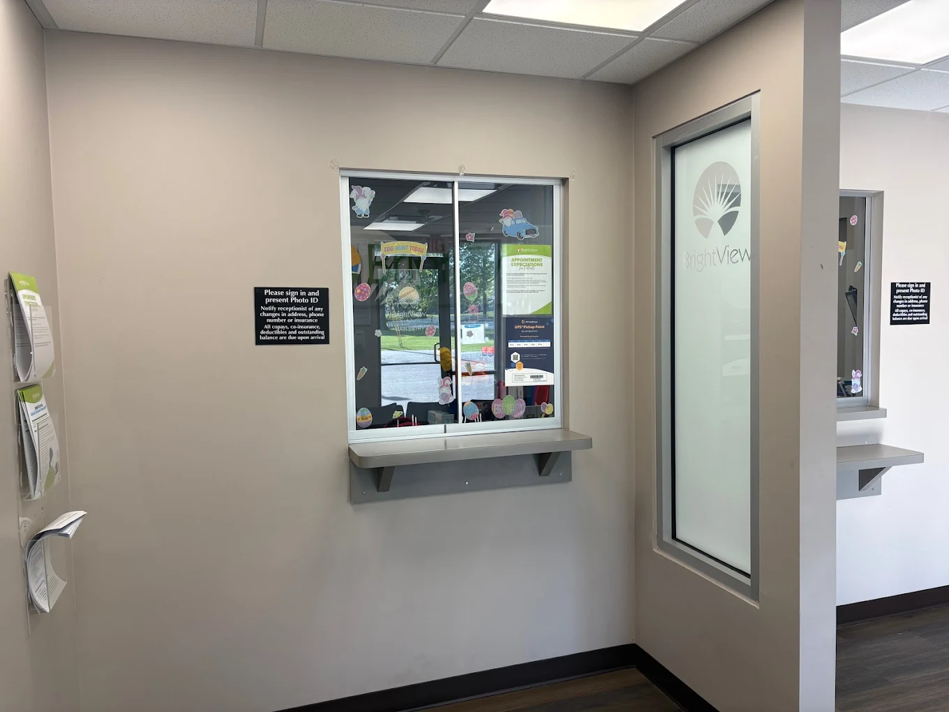 BrightView Health check-in area with glass service window and counter, informational signs, brochure holders, and frosted window displaying the BrightView logo