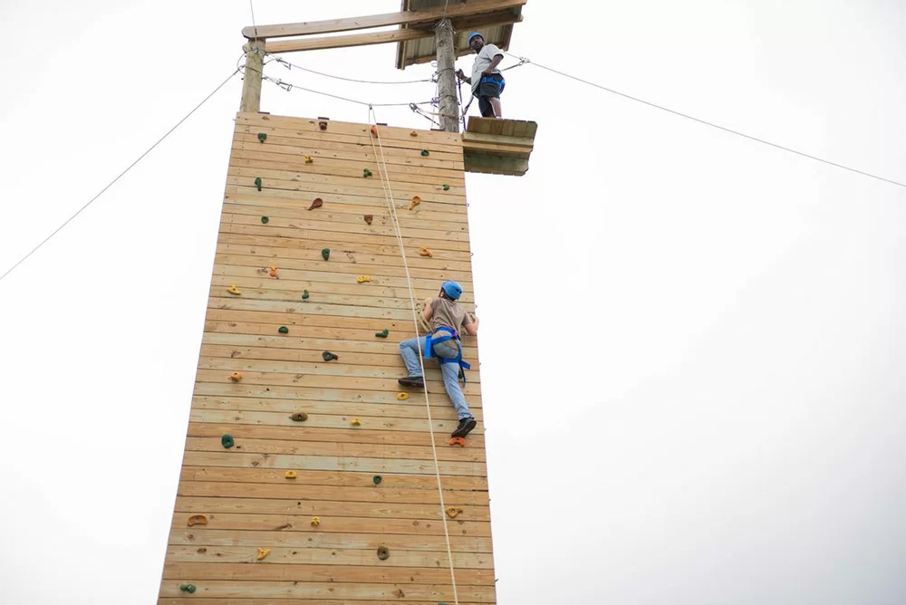 Person climbing tall wood rock wall with safety harness
