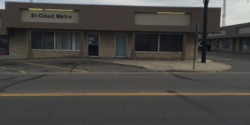 A modest building with a "St Cloud Metro" sign, large windows, and a small entrance, situated along a quiet street under an overcast sky. 