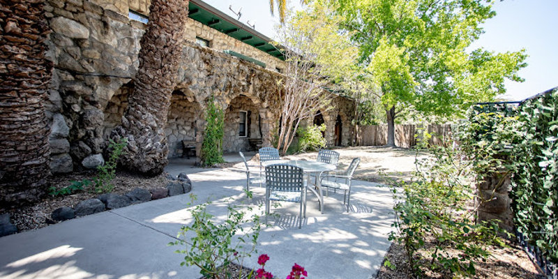  A peaceful outdoor patio with shaded seating, surrounded by greenery, stone arches, and bright flowers, offering a tranquil spot to relax.