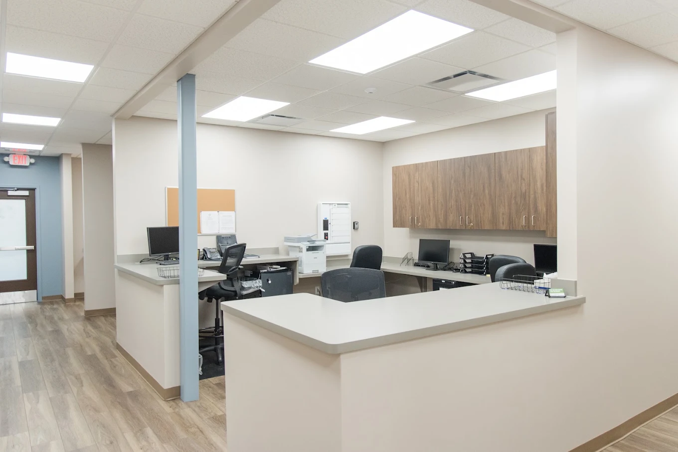 Open staff workstation with reception counters, computers, office chairs, printer, and overhead wood cabinets