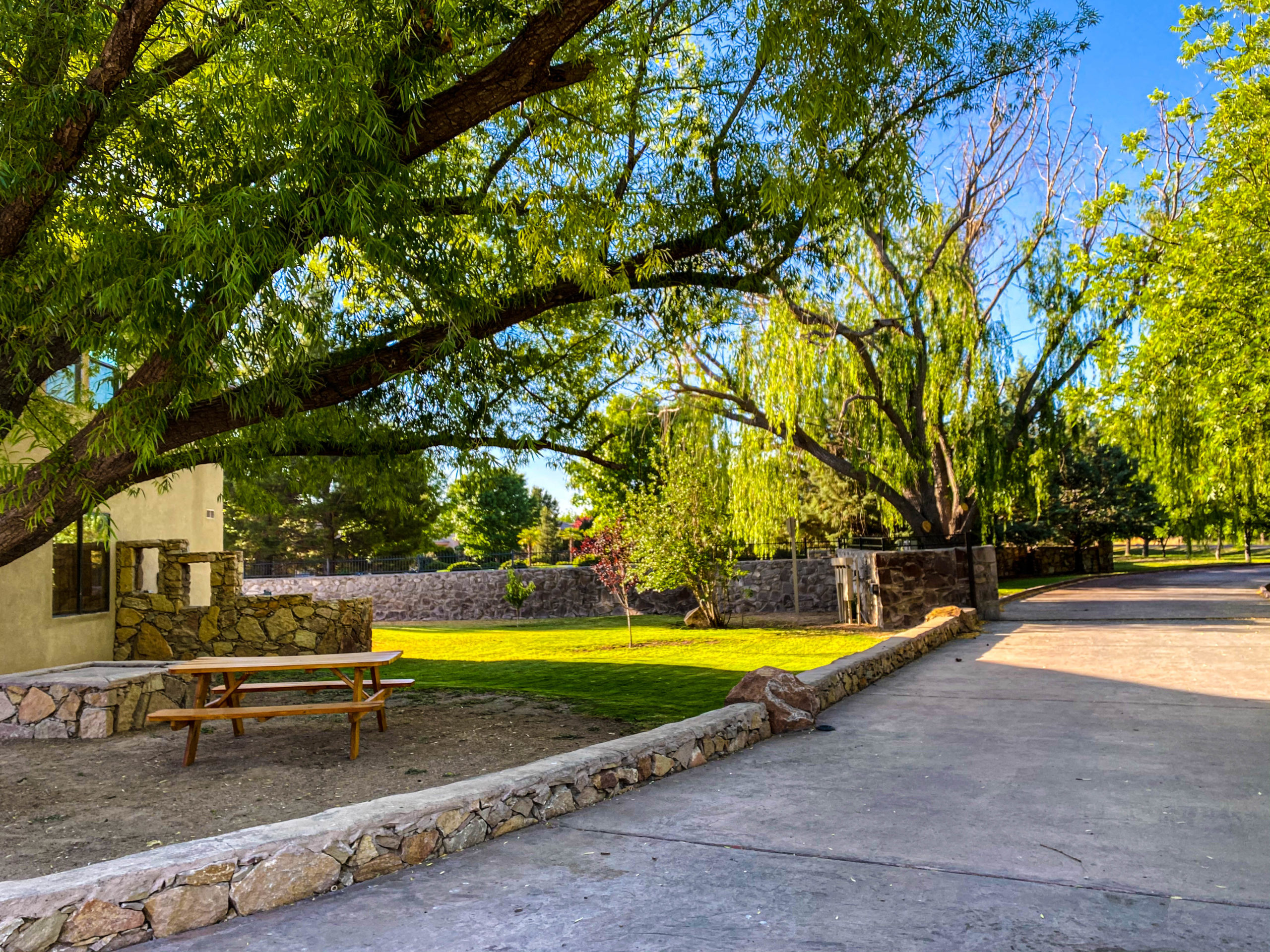 Tree-lined outdoor area with stone accents, bench-style seating, lawn, and surrounding greenery along a concrete path