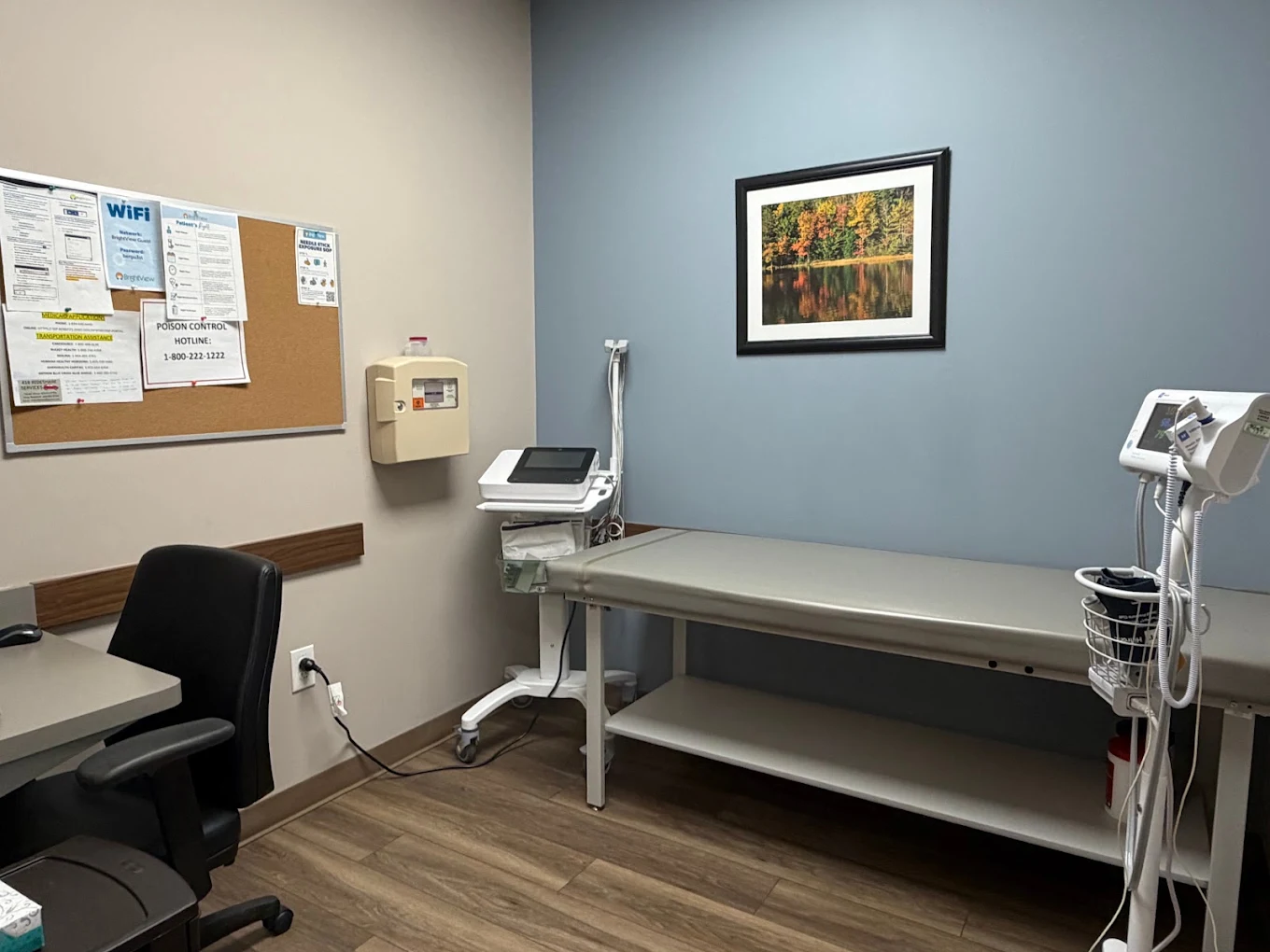 Clinical exam room at BrightView Health Mansfield with an examination table, medical monitoring equipment, office chair, bulletin board, and blue accent wall