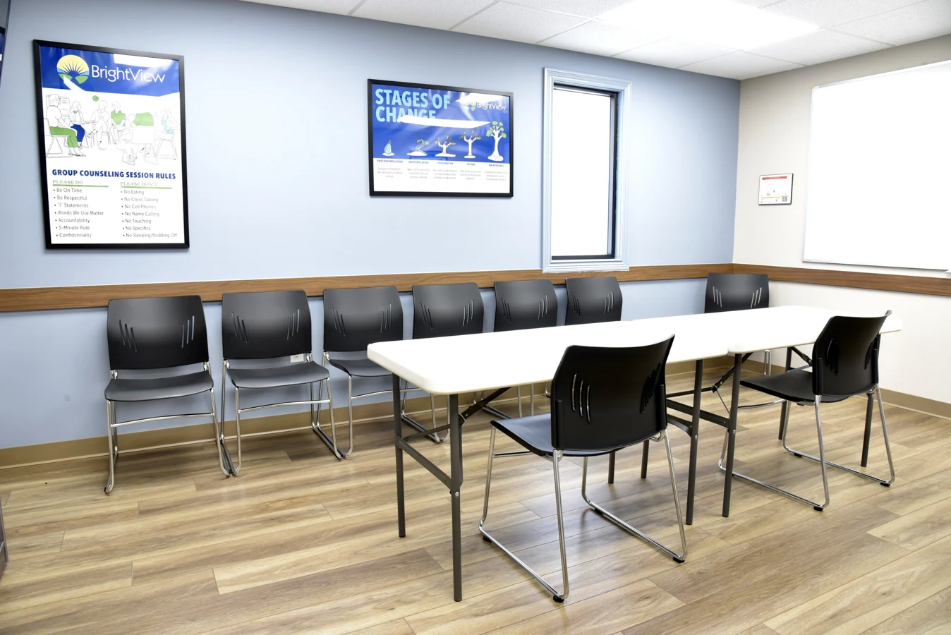BrightView Health group counseling room with white tables, black chairs, wood-look flooring, blue walls, whiteboard, and framed recovery and counseling posters