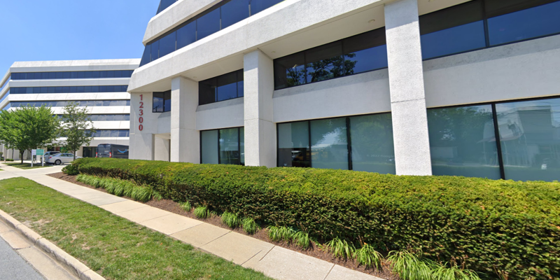  Modern office building with large reflective windows, white exterior, and well-manicured greenery lining the walkway, creating a clean, professional look.