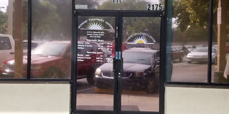 Glass door with "Sunrise Treatment Center" signage, reflecting cars parked outside. The center’s operating hours and services are listed.