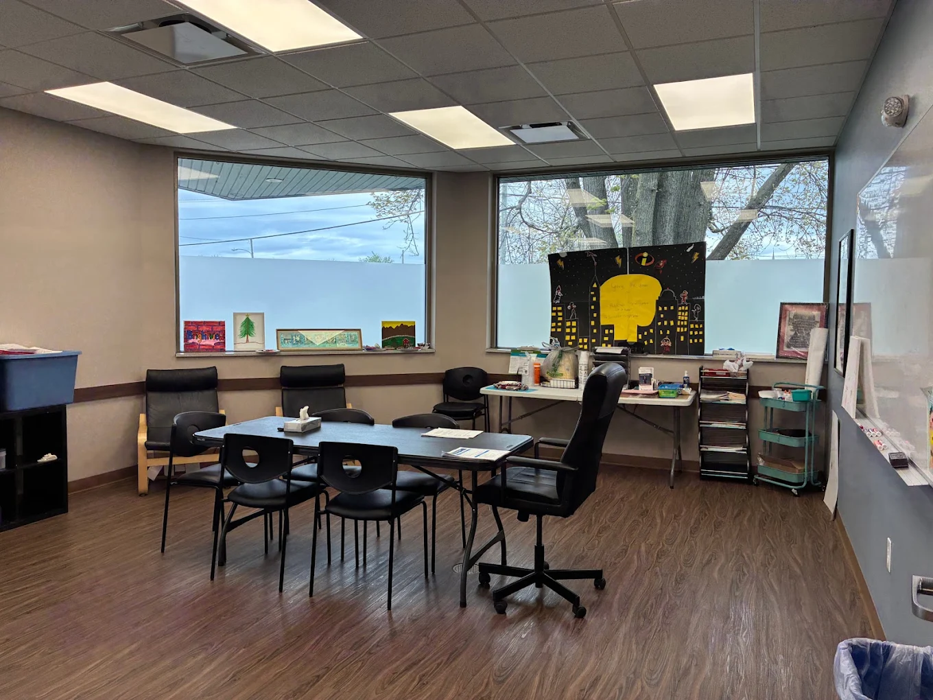 Group counseling room with tables, chairs, large windows, whiteboard, storage shelves, and activity materials