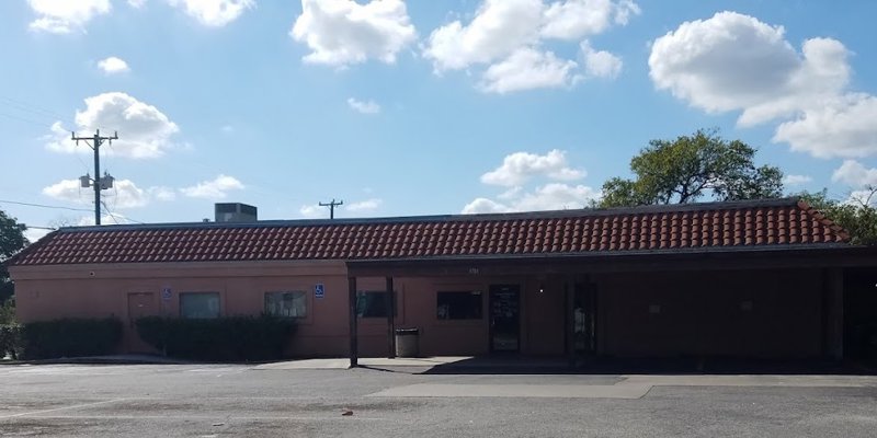 A building with a red-tiled roof and a spacious parking lot, bathed in sunlight under a bright blue sky with fluffy clouds. Peaceful surroundings.