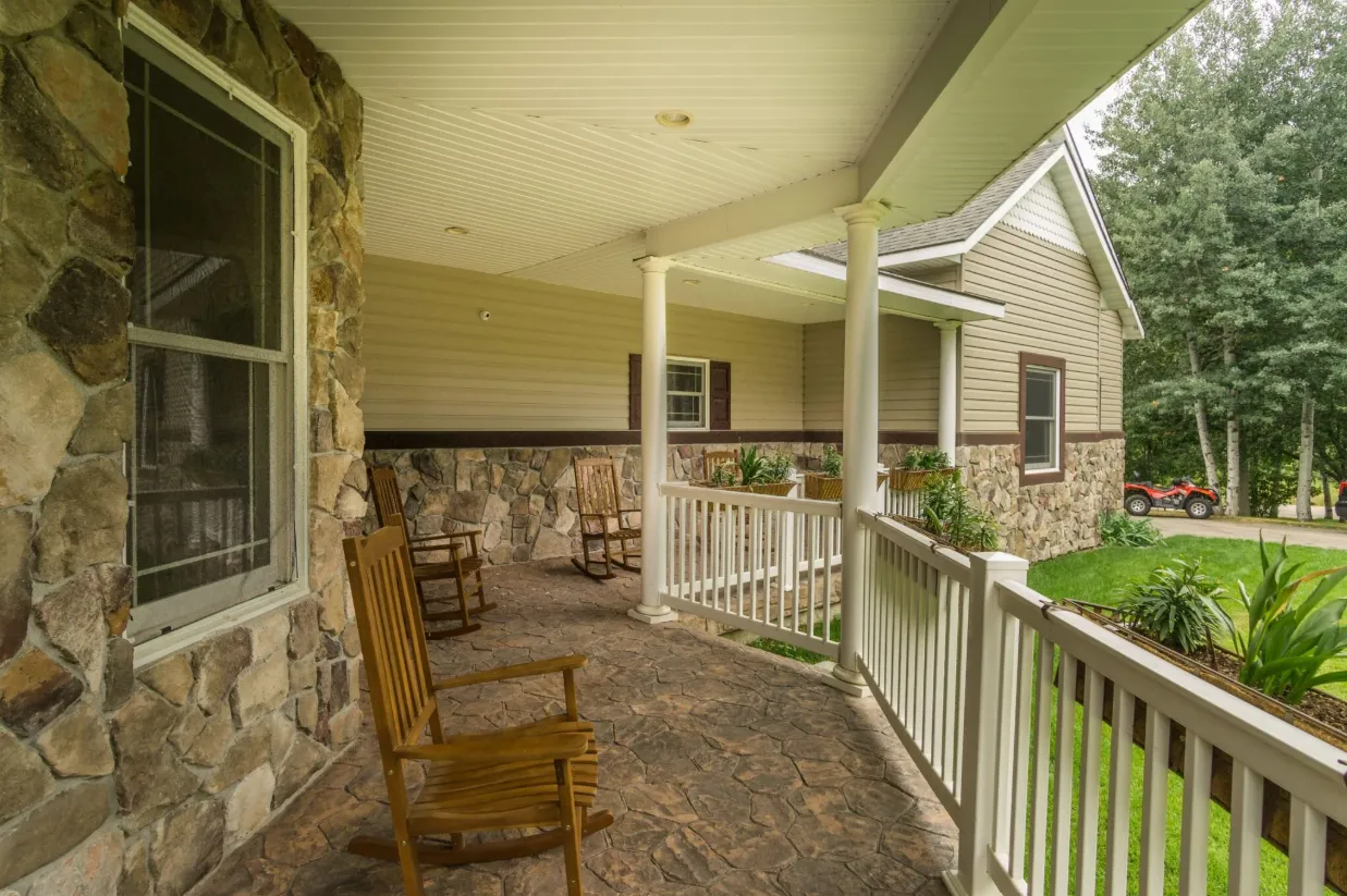 Covered porch with stone siding, white railing, rocking chairs, and view of the lawn and trees